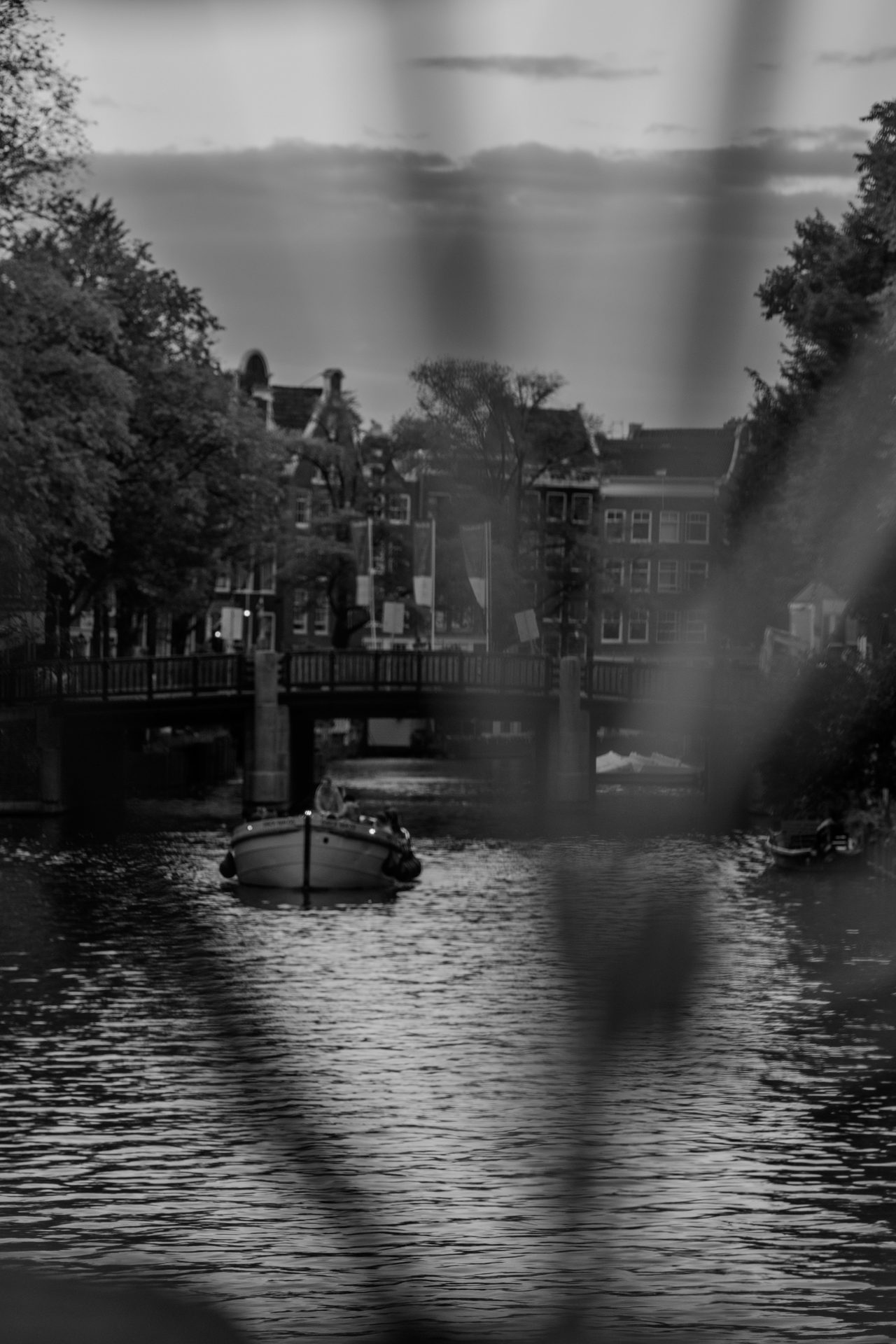 Boat heading up the canal toward a bridge, seen through the blurred bars of a railing, black and white