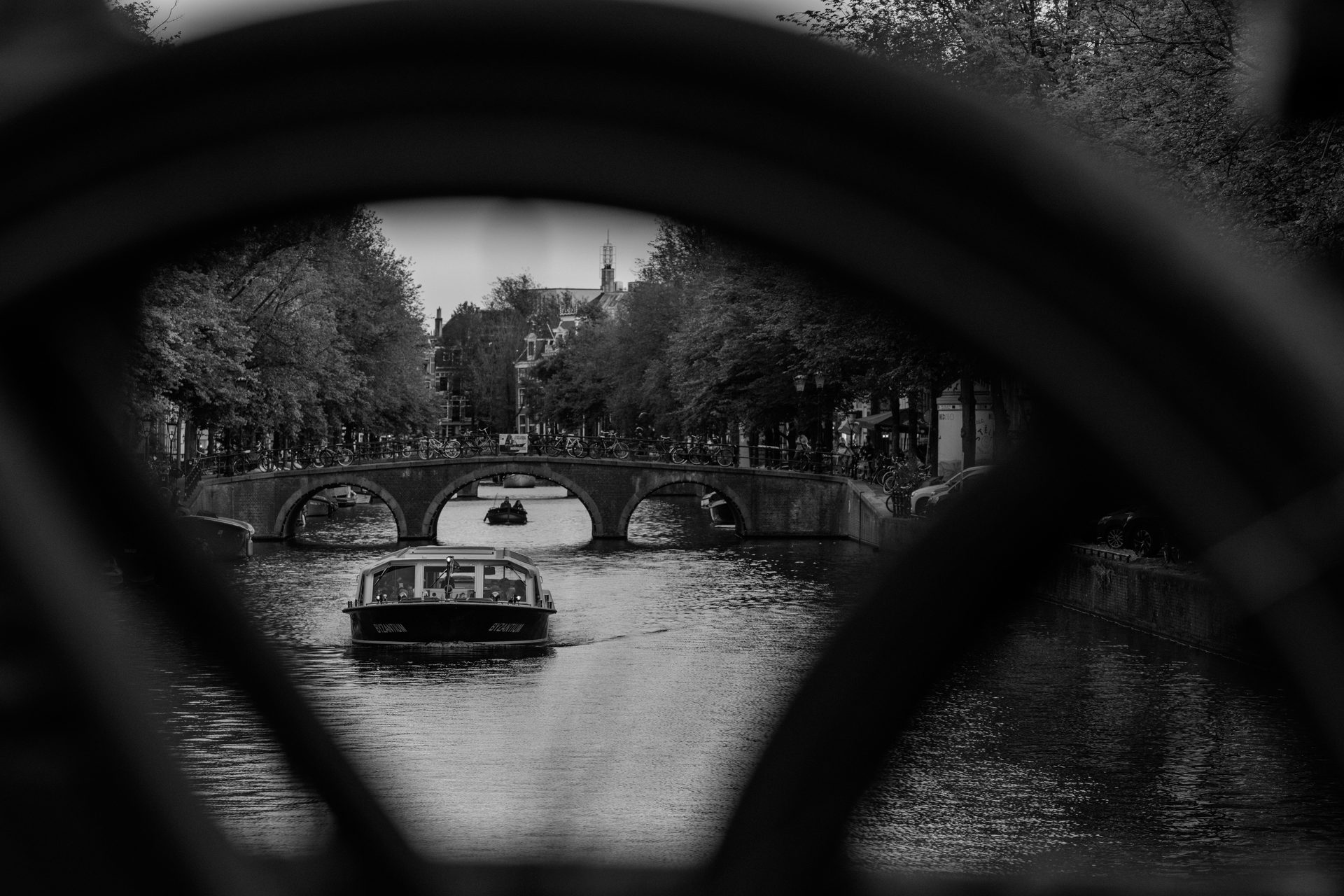Boat passing under a three-arch bridge, framed inside a bicycle wheel, black and white