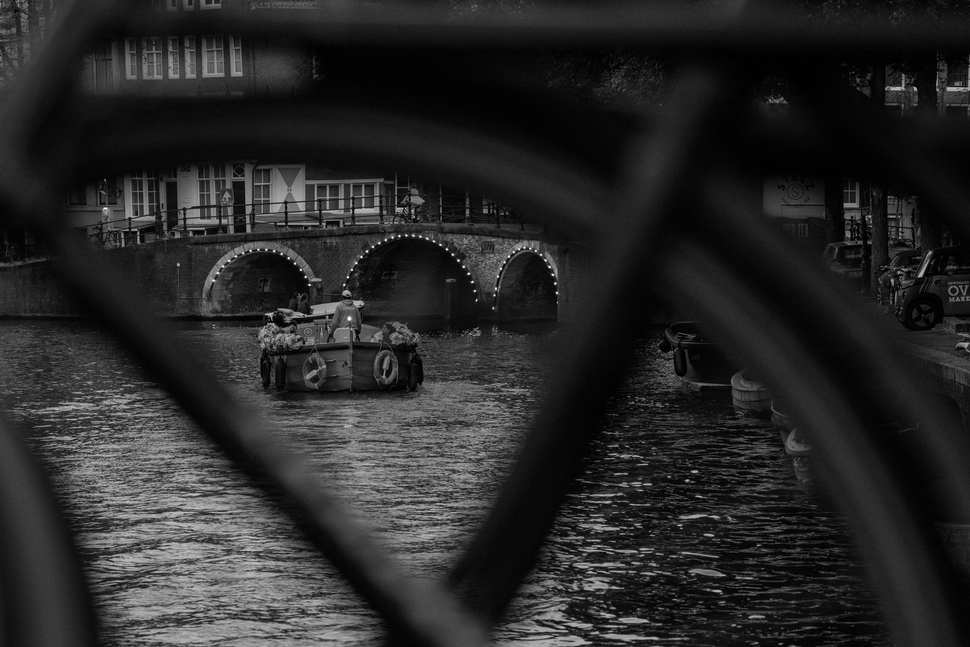 Boat loaded with flowers under a brick bridge lit with bulbs, seen through a bicycle frame, black and white