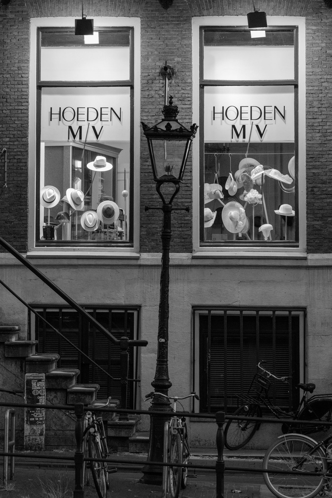 Two lit hat-shop windows, white hats suspended, a streetlamp between them, black and white