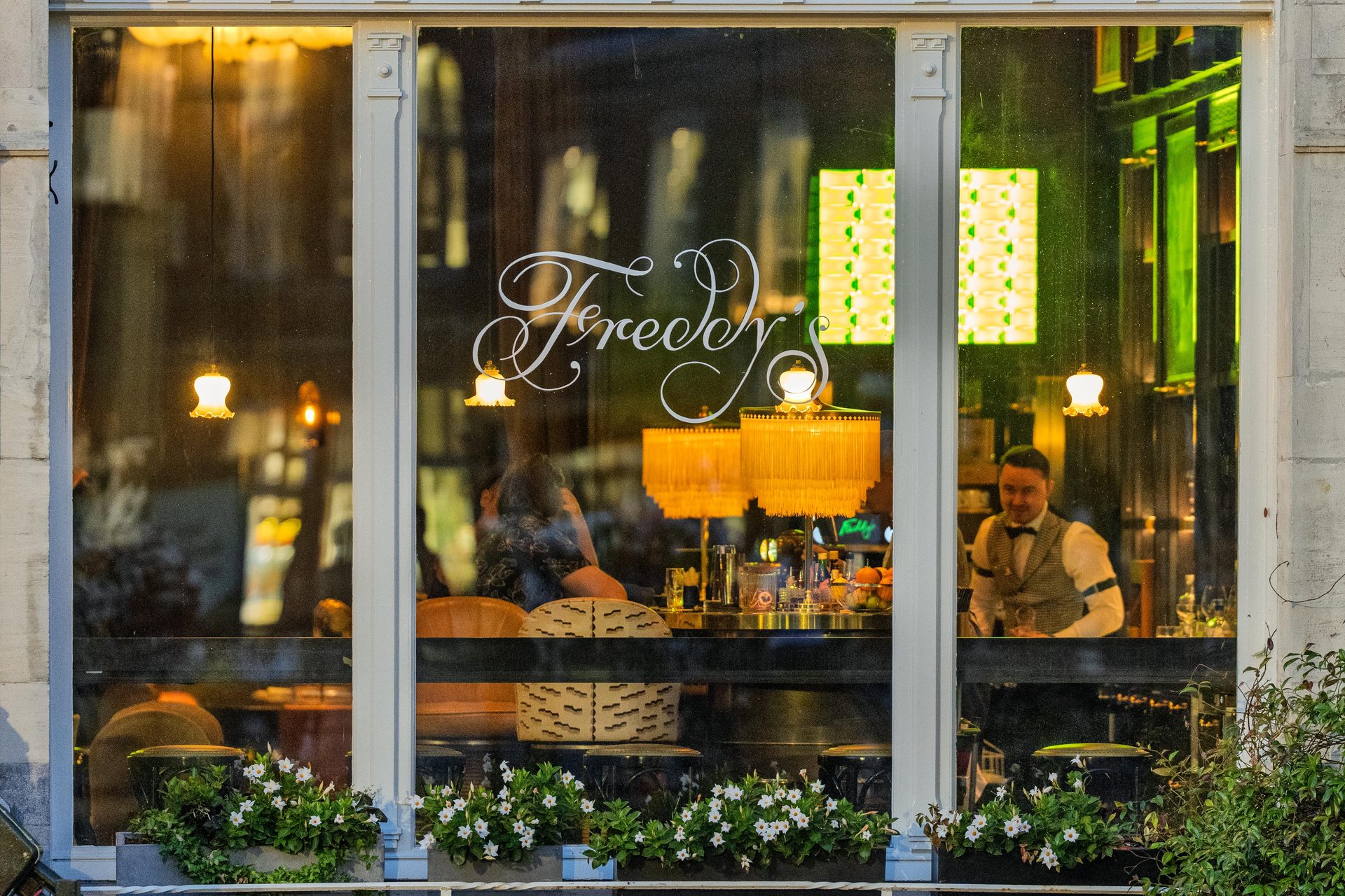 Bartender behind a lit bar window, fringed lamps and green neon, seen from the street