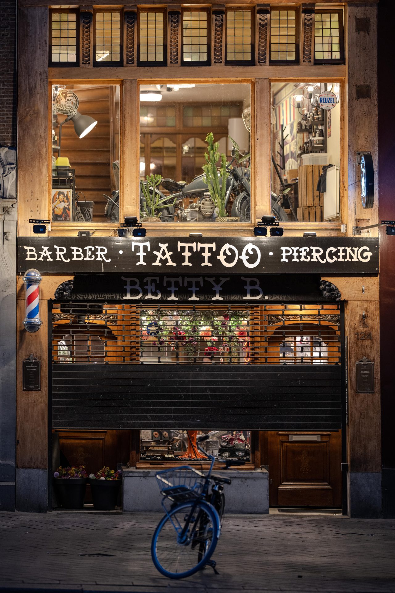 Barber and tattoo shopfront at night, shutter half down, blue bicycle parked outside