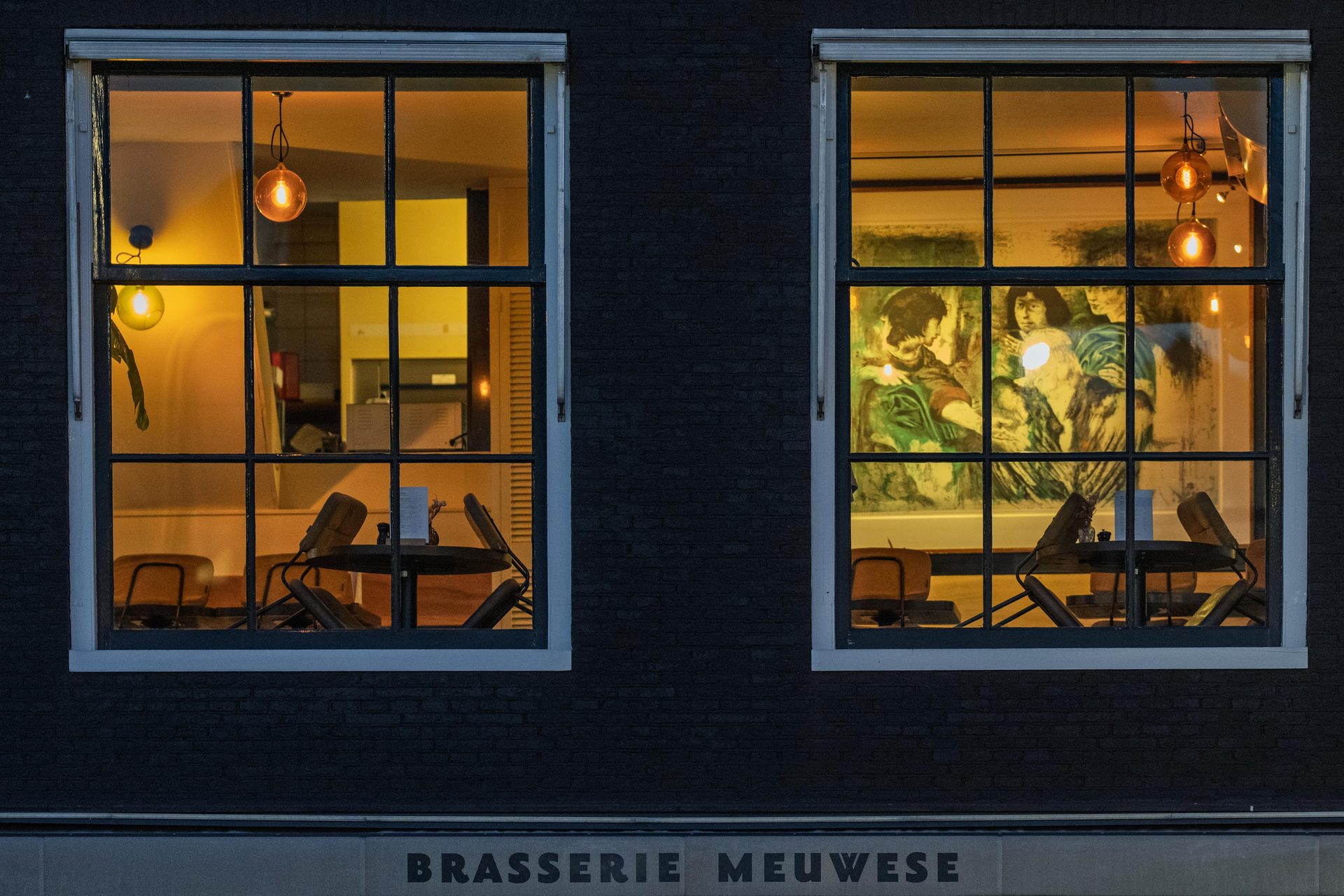 Two lit brasserie windows in a black brick façade, laid tables and a large painting inside
