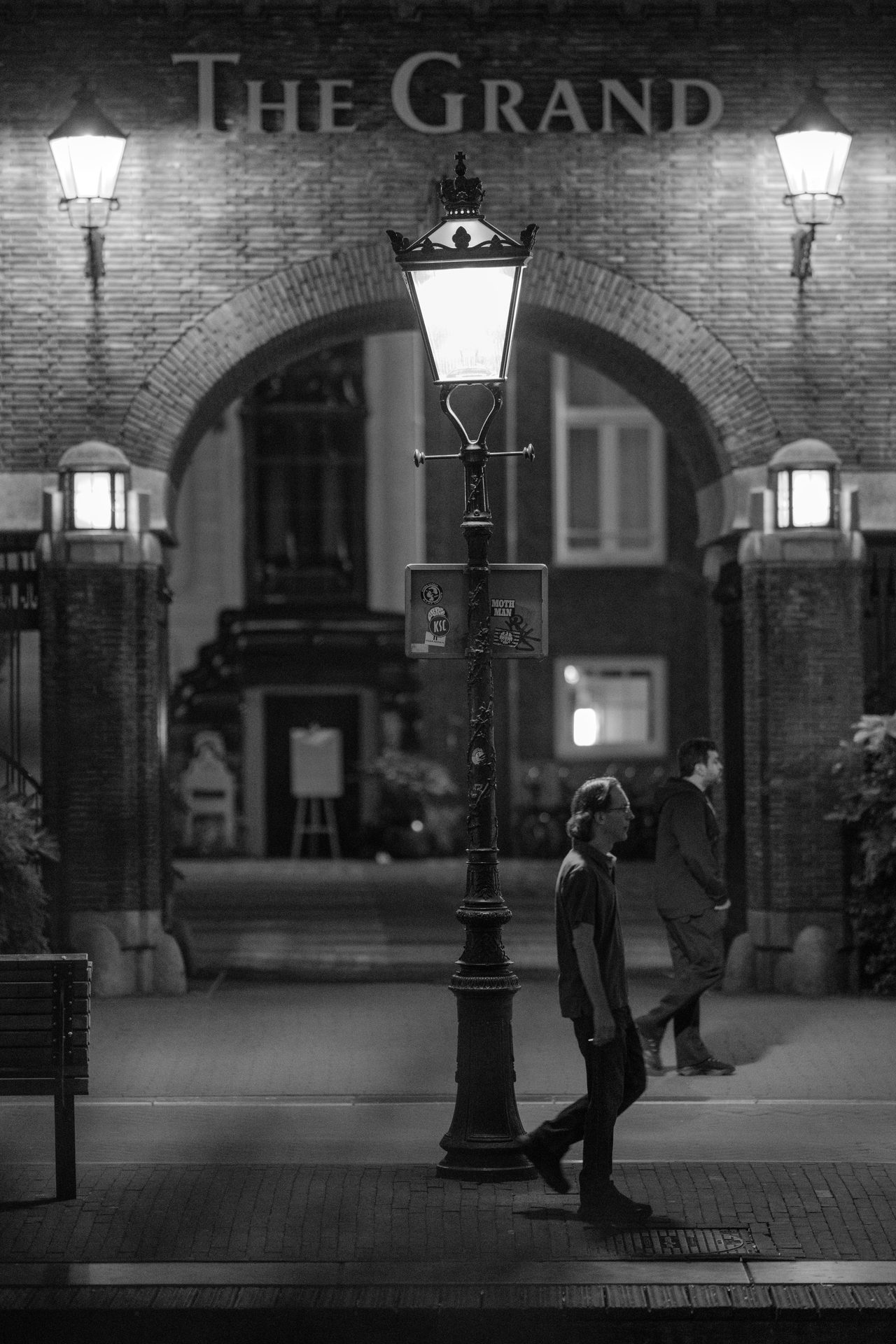 Old streetlamp in front of the arched entrance of The Grand hotel, two passers-by walking opposite ways, black and white