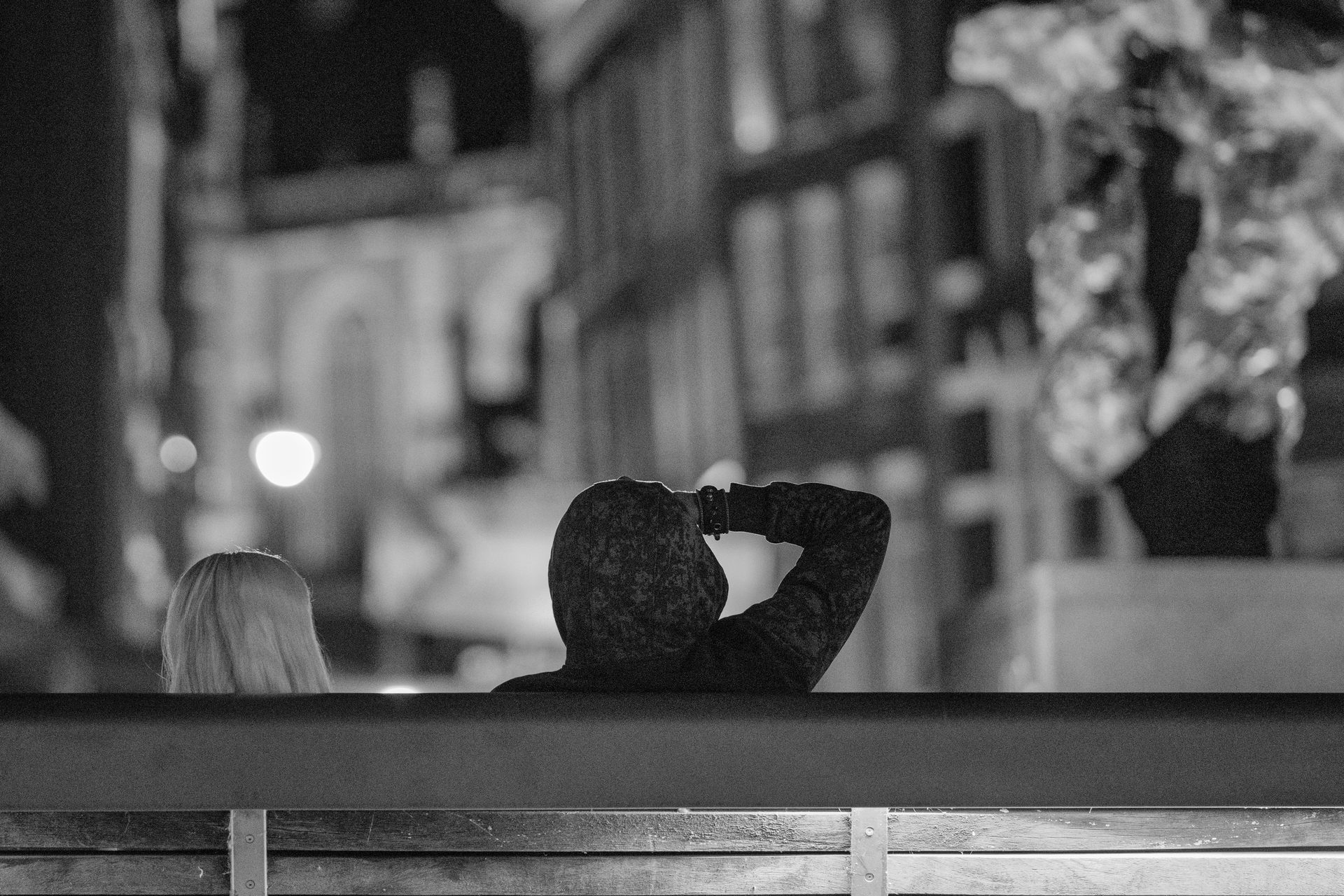 Two people from behind on a canal-side bench, one arm folded behind a head, blurred façades beyond, black and white