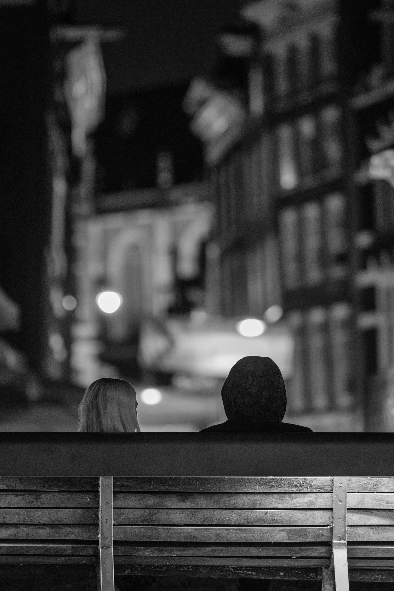 Two figures sitting side by side on a bench facing the canal, blurred street lights beyond, black and white