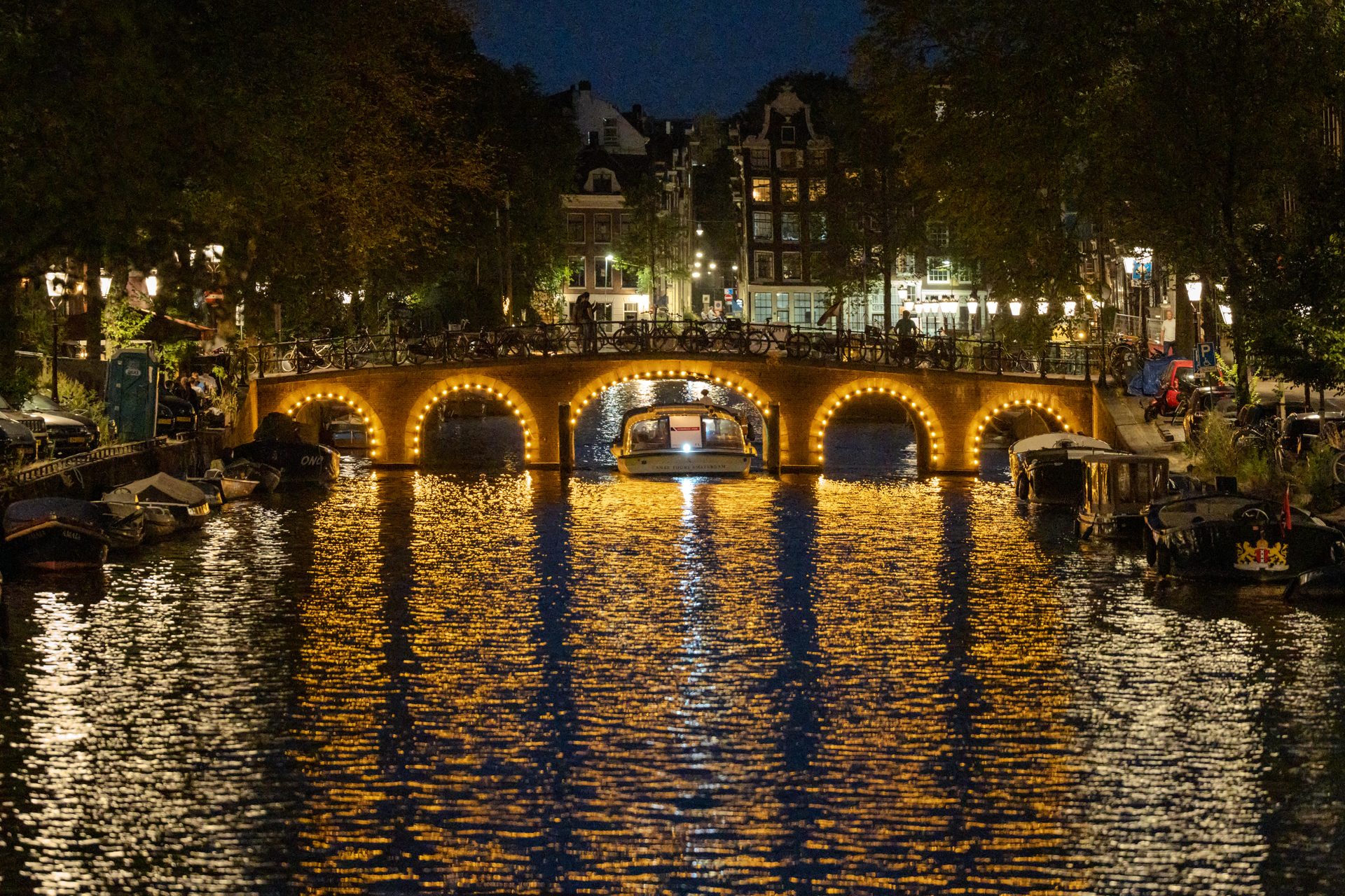 Reguliersgracht bridge with bulb-lit arches, a boat passing beneath, golden reflections on the water