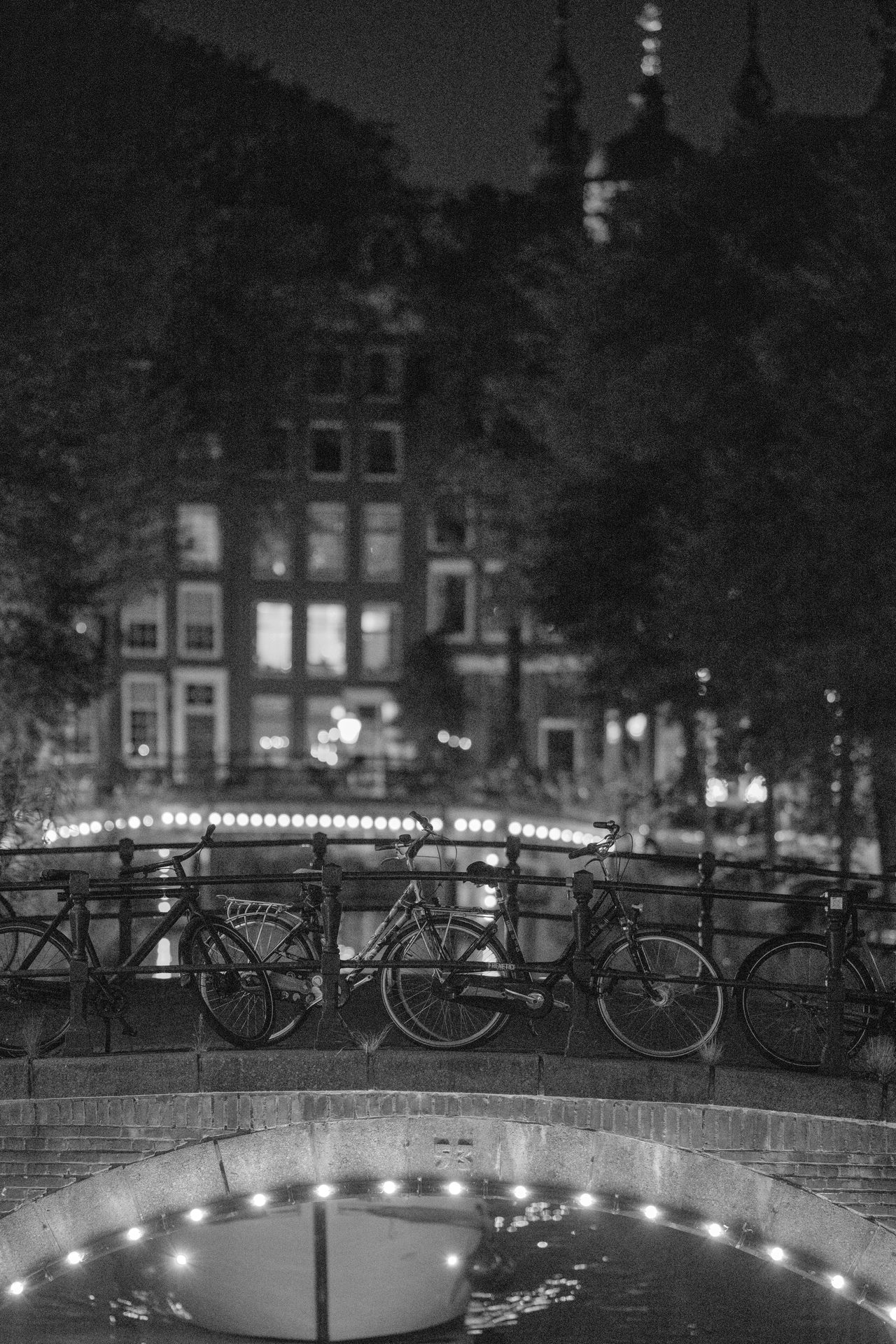 Bicycles lined along a canal bridge railing, a bulb-lit arch in the foreground, black and white