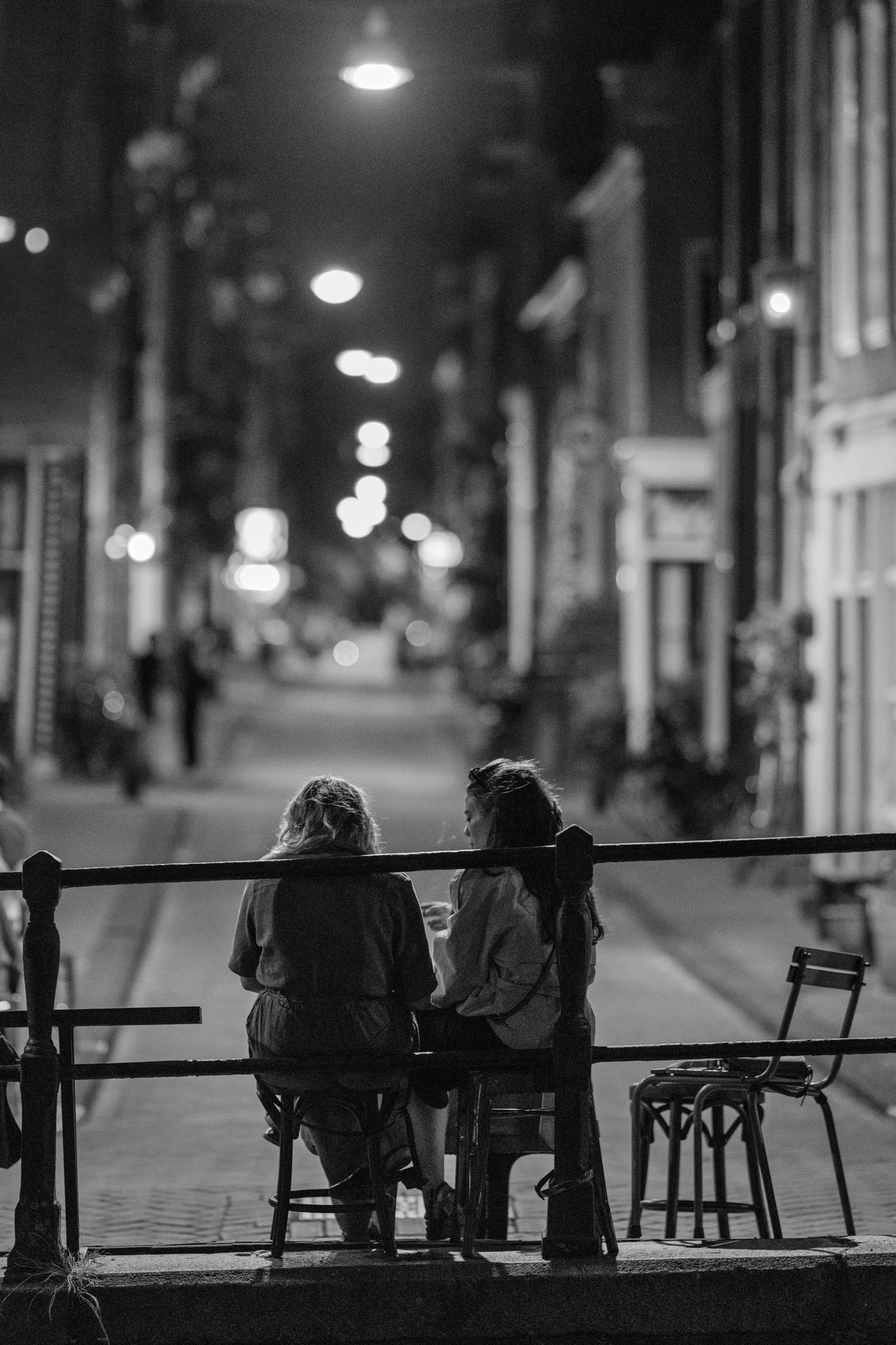Two women at a canal-side terrace table, empty street and a receding line of lamps beyond, black and white